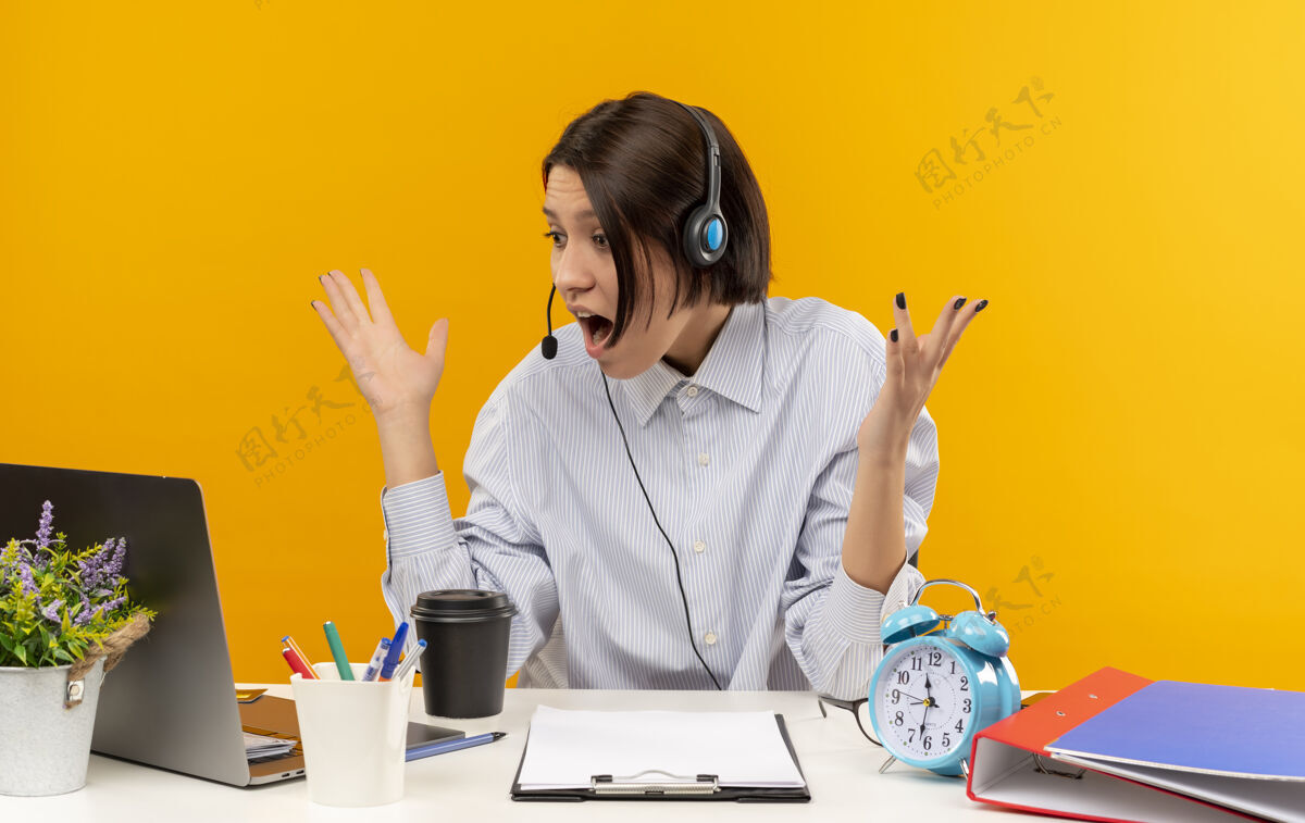 耳机印象深刻的年轻呼叫中心的女孩戴着耳机坐在办公桌旁举手 看着笔记本电脑上孤立的橙色坐着笔记本电脑女孩