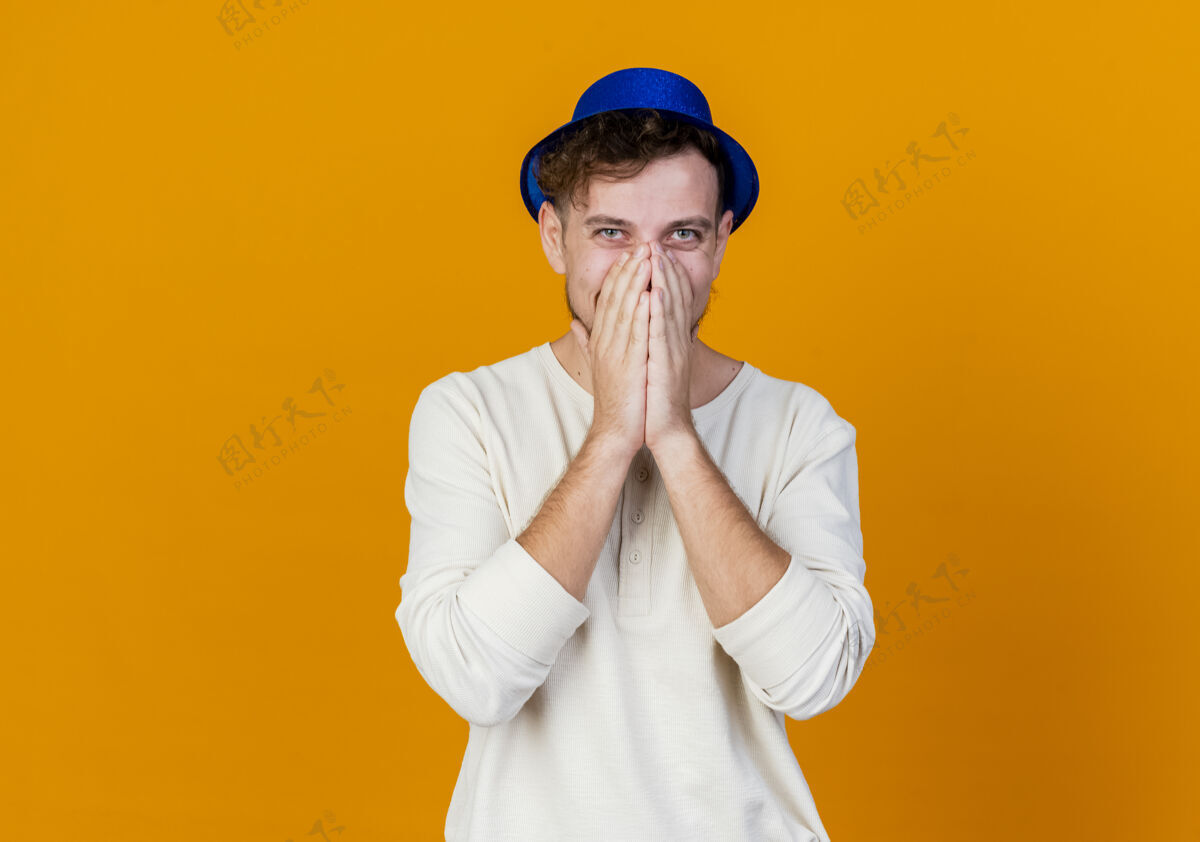 相机面带微笑的年轻英俊的斯拉夫党人戴着党的帽子看着相机保持手放在嘴上橙色背景与复制空间隔离嘴巴派对微笑