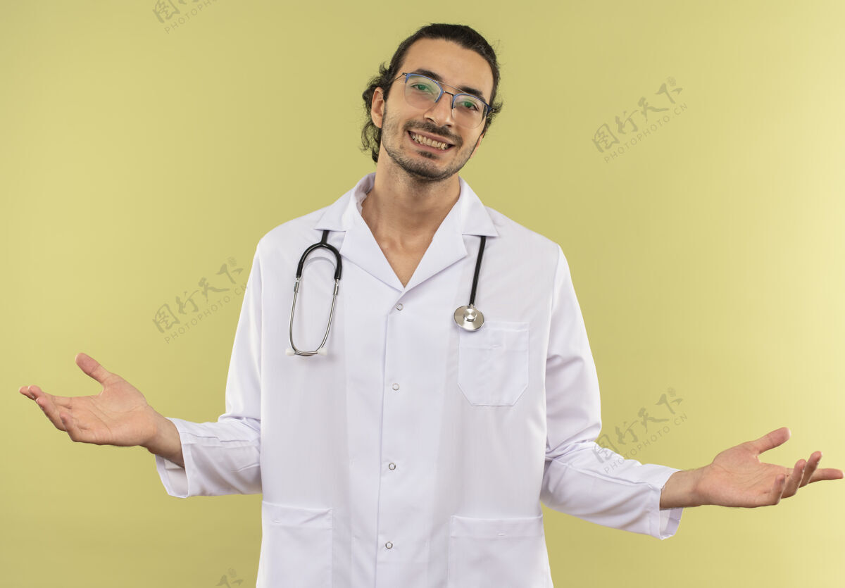 光学微笑的年轻男医生 戴着眼镜 穿着白色长袍 带着听诊器 在隔离的绿色墙壁上摊开双手 留着复印空间穿眼镜微笑