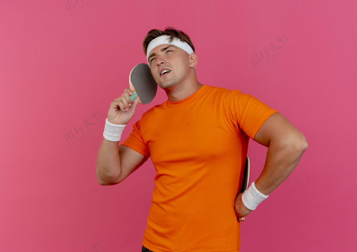 自信自信的年轻帅气的运动型男人戴着头带和腕带 拿着乒乓球拍 假装在电话里说话 粉红色的地板上有一个复制空间穿太空球拍