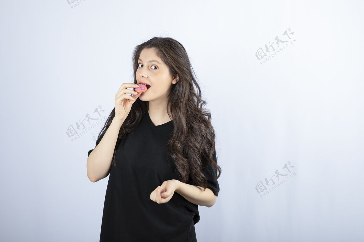站着穿黑衣服的年轻女孩在白墙上吃粉红色的饼干女孩甜点女人