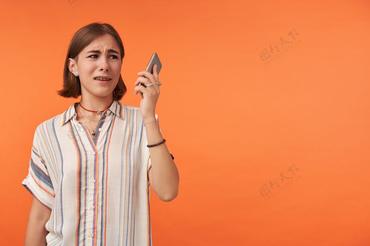 悲伤不开心的女孩拿着智能手机看穿着条纹衬衫 戴着牙套和手镯看着右边 在橙色的墙上复制空间时尚电话牙齿