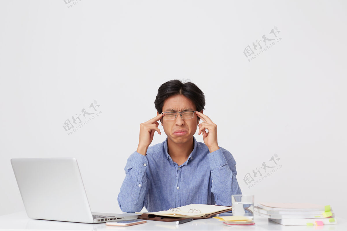 压力一个戴着眼镜 眼睛闭着摸太阳穴 心情紧张 心情不愉快的亚洲年轻商人 在白墙上的桌子旁用笔记本电脑感到压力封闭互联网笔记本电脑
