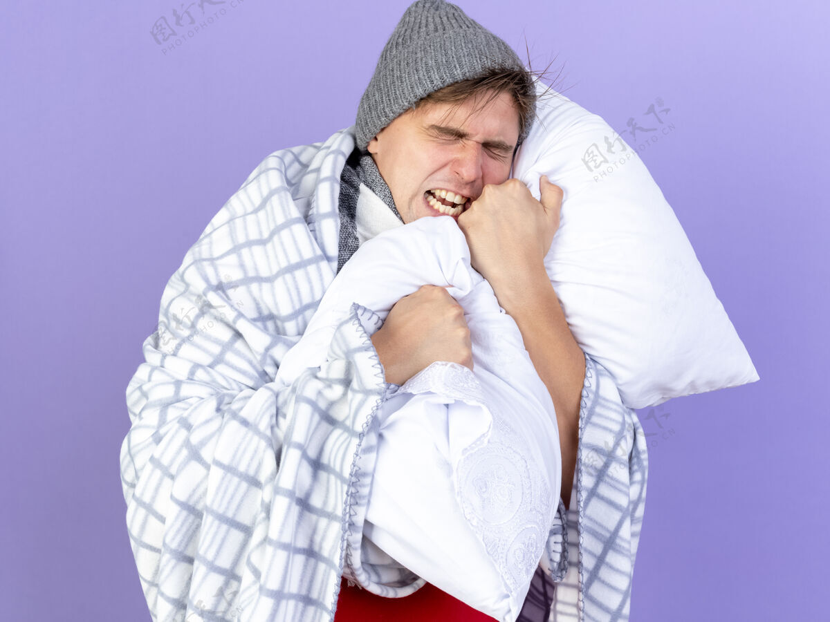尖叫紧张的年轻英俊的金发病男子戴着冬天的帽子和围巾裹在格子布抱着枕头尖叫着闭着眼睛孤立在紫色的背景封闭压力眼睛