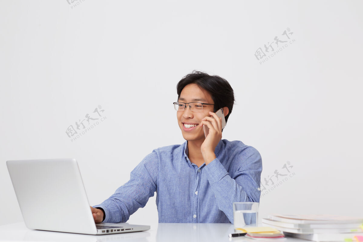 笔记本电脑快乐成功的亚洲年轻商人戴着眼镜 穿着蓝色衬衫 在白墙上的桌子旁用笔记本电脑打电话快乐手机手机