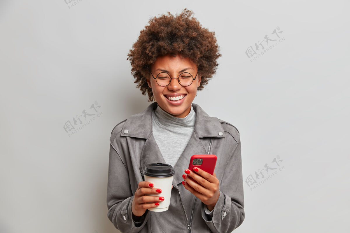 夹克快乐乐观的女人在室内开怀大笑 喝咖啡去 手里拿着手机 等着有人 白天有散步 表达积极的情绪正面牙关拿出来