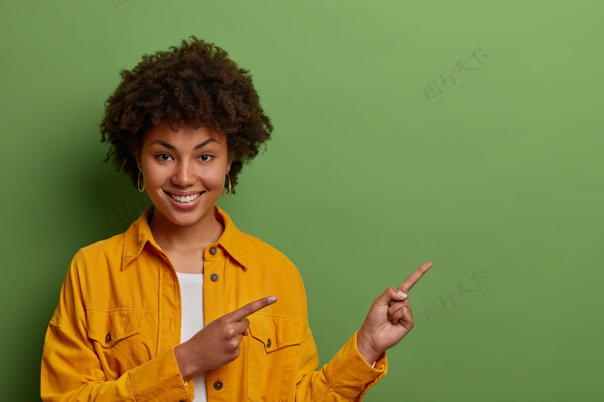 年轻可爱的黑皮肤女人 非洲式头发 手指指向右边 建议朝这个方向走 展示很棒的产品 穿黄色夹克 室内点模型