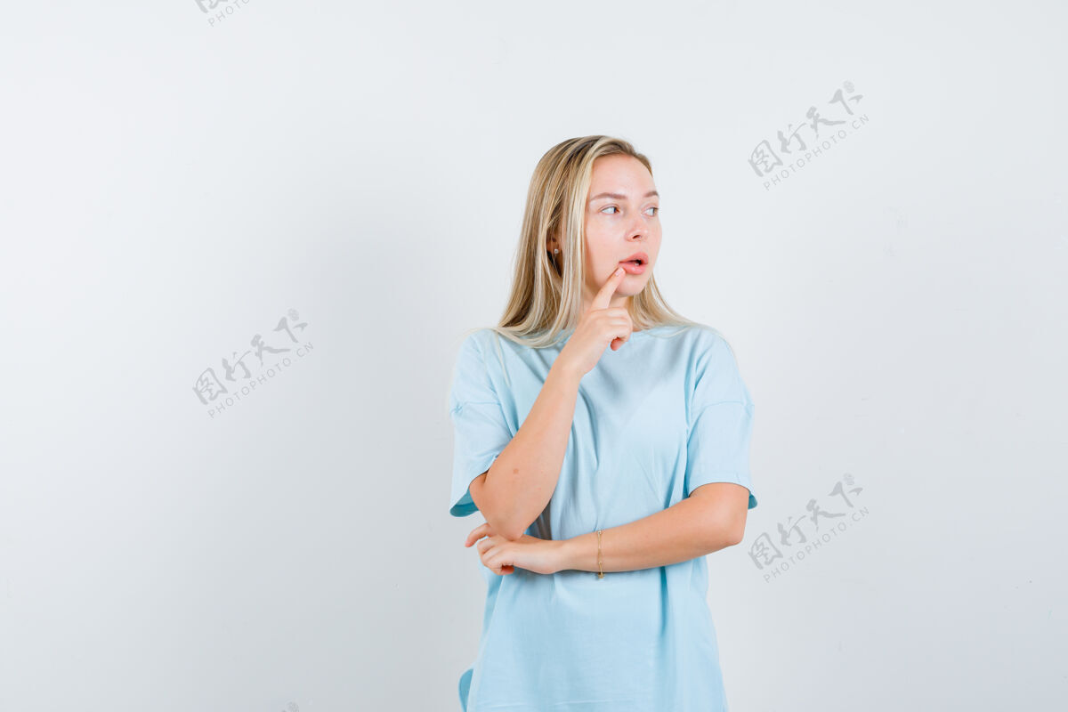 嘴巴穿着蓝色t恤的金发女孩站在思考的姿势 把食指放在嘴边 看起来沉思 前视图年轻模特微笑