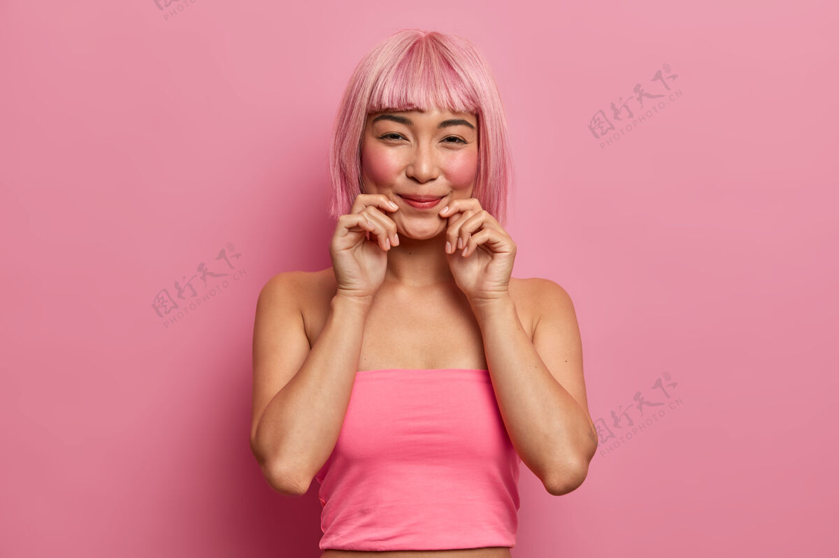 胭脂美丽的粉红头发的亚洲女士微笑愉快 双手紧握嘴唇 听到喜讯高兴 身穿玫瑰色上衣站在室内单色民族女孩用时髦的发型表达良好的情感模特积极室内