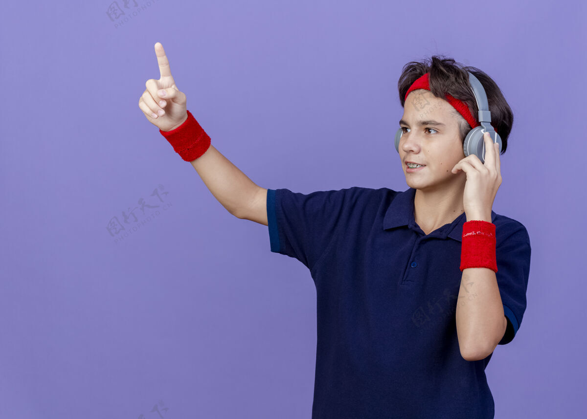 耳机高兴的年轻帅气的运动男孩戴着头带和护腕 戴着戴着牙套的耳机看着侧面触摸耳机指向紫色背景上的复制空间孤立腕带运动佩戴