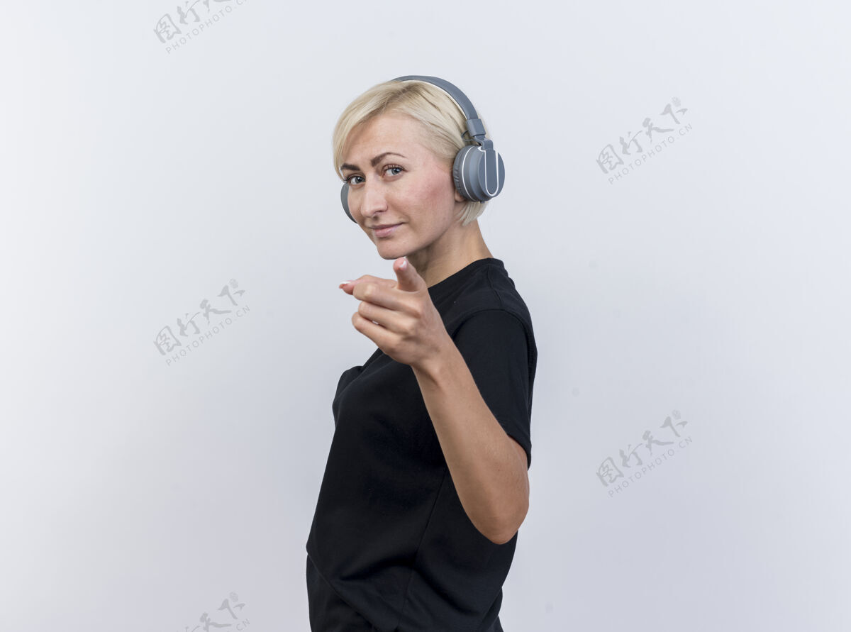 空间自信的中年金发斯拉夫女人戴着耳机站在侧视图中看着并指着隔离在白色背景上的相机 还有复印空间指向中年复制