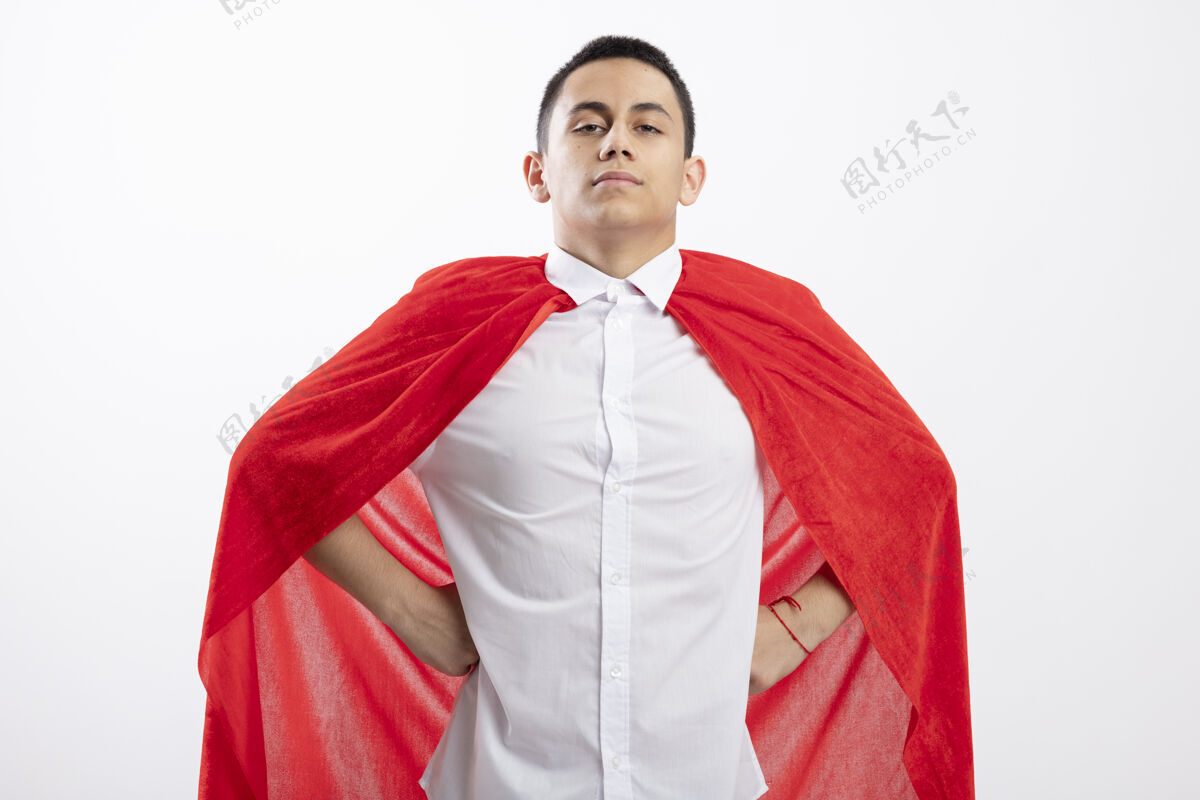 年轻穿着红色斗篷的自信的年轻超级英雄男孩手放在腰上看着白色背景上的相机自信腰手