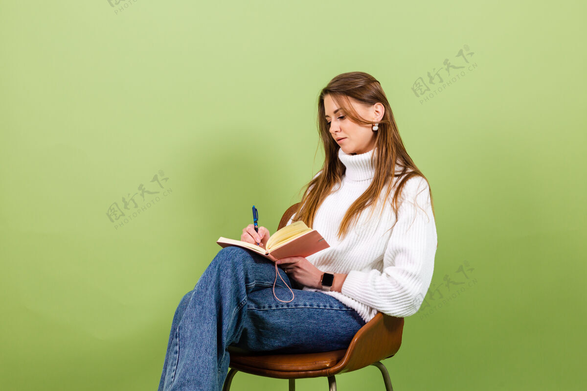 微笑漂亮的欧洲女人穿着休闲的白色毛衣孤立 可爱快乐的坐在椅子上用记事本和笔狡猾美丽学生