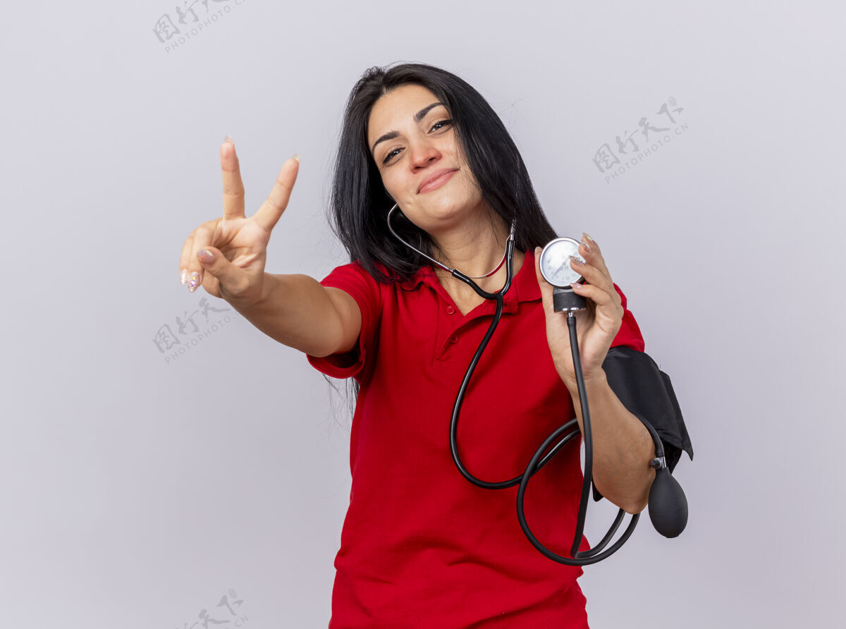 压力戴着听诊器的年轻白种人病女孩高兴地看着摄像机 用血压计测量她的血压 在白色背景上做着隔离的和平标志和平听诊器佩戴