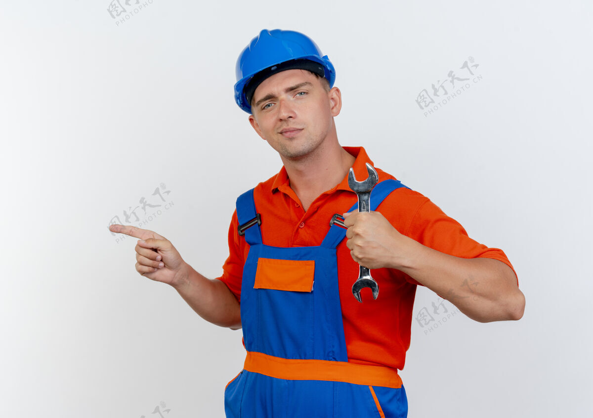 安全高兴的是年轻的男建筑工人穿着制服 戴着安全帽 手持扳手 指着旁边的白色制服壁板请
