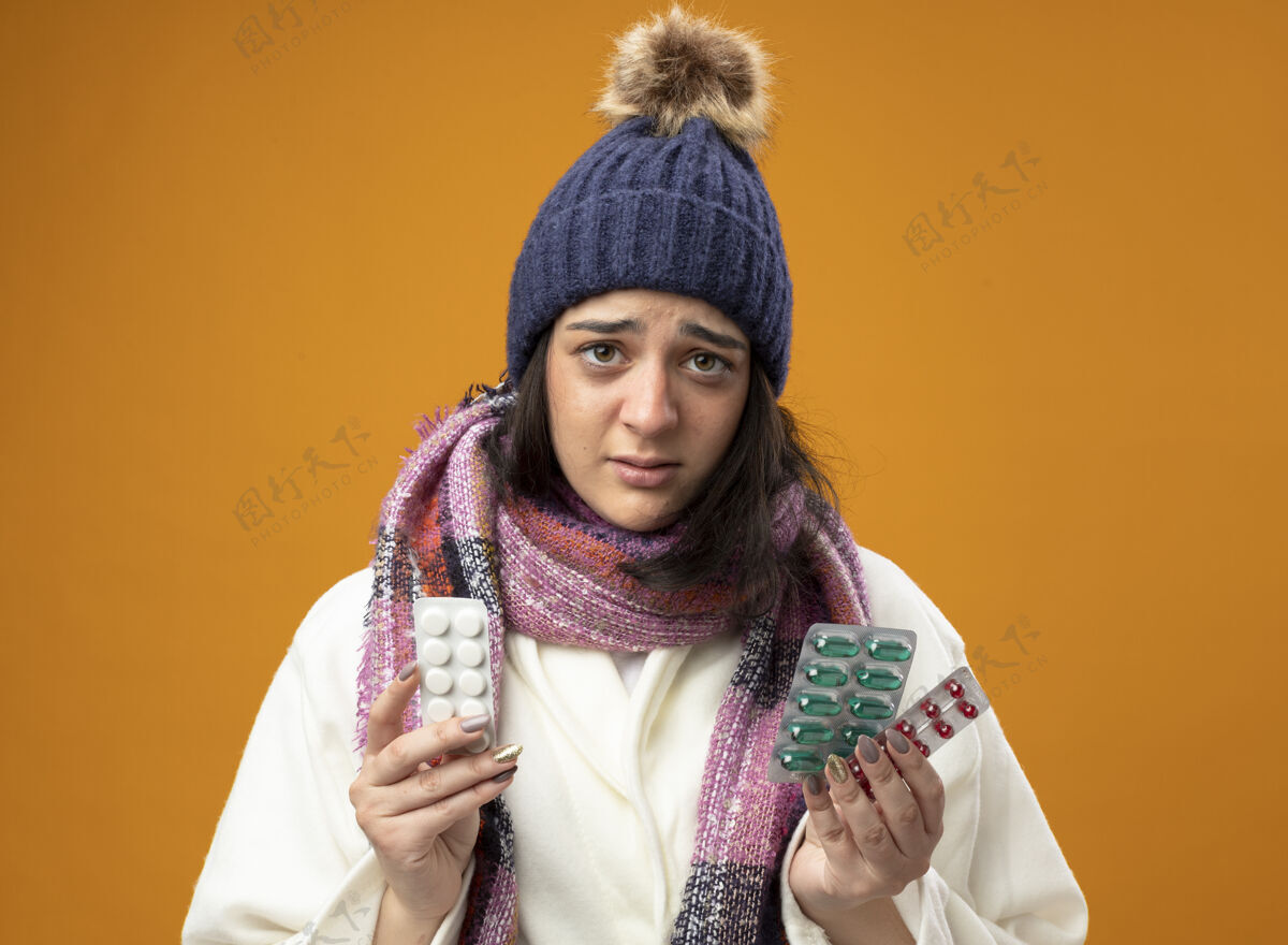 冬天印象深刻的年轻生病的女人穿着长袍 戴着冬天的帽子和围巾 拿着一包包的药丸 看着前面孤立的橙色墙壁药丸感觉疾病