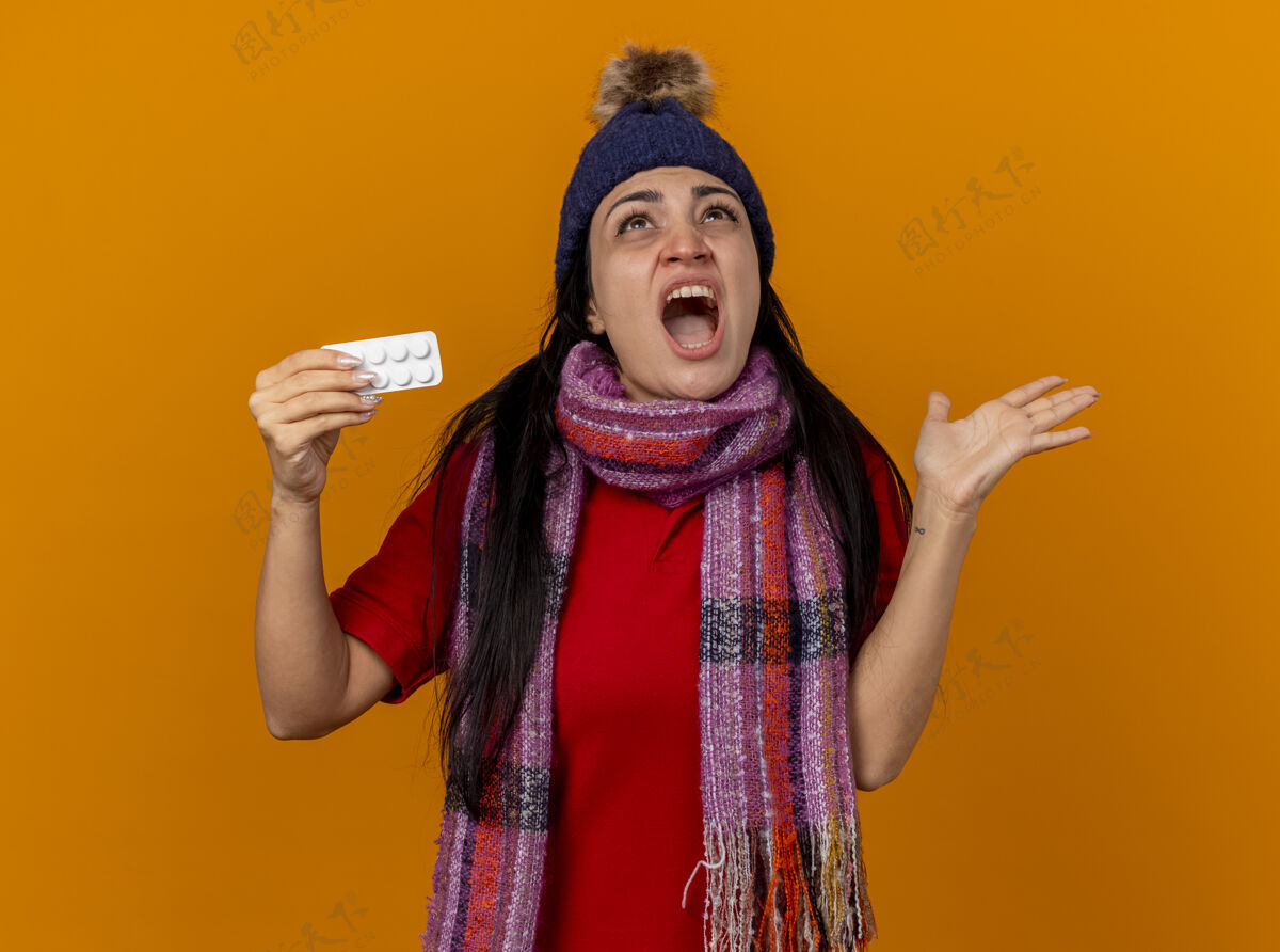 空愤怒的年轻生病的女人戴着冬天的帽子 戴着围巾 手里拿着一包药片 抬起头来 手上空空如也 在橙色的墙上尖叫着表情女人冬天