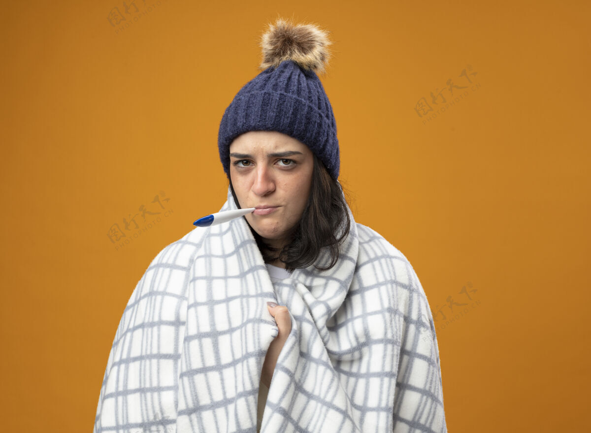 年轻人皱着眉头的年轻病妇 戴着格子布裹着的长袍冬帽 抓着格子布看着前面 嘴里叼着温度计 隔离在橙色的墙上人疾病抓住