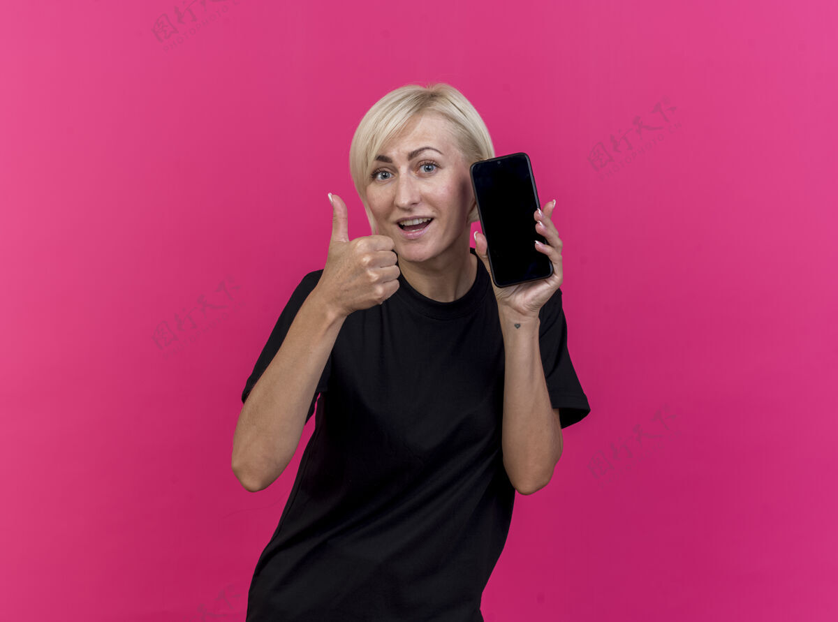 感觉令人印象深刻的中年金发女子显示手机和拇指向上看着前面孤立的粉红色墙上拇指人市民
