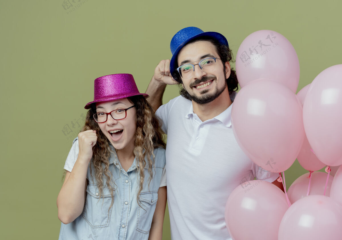 年轻一对微笑的年轻夫妇戴着粉色和蓝色的帽子站在气球旁边 在橄榄绿上显示“是”的手势帽子站立情侣