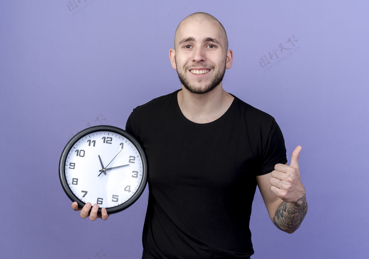 紫色面带微笑的年轻人举着挂钟 他的大拇指孤立地竖在紫色的墙上墙壁抱着时钟