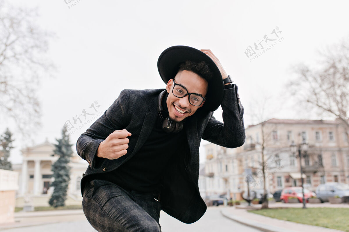 放松穿着考究的非洲男人微笑着在城市广场上跳舞在灰暗的天空下 一个笑嘻嘻的黑人摸着他的帽子在户外摆姿势寒冷帽子城市景观