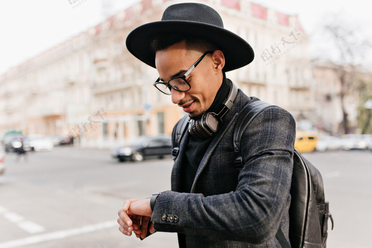 城市穿着考究的非洲绅士在看他的手表户外照片中的混血儿戴着帽子 背着皮包配件街道男人
