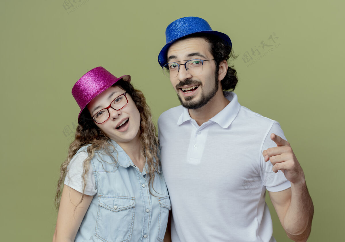 橄榄色微笑的年轻夫妇戴着粉色和蓝色的帽子 男人在橄榄绿上向你展示孤立的姿势穿着绿色帽子