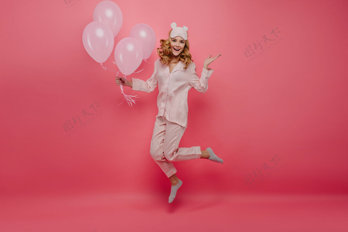 气球穿着袜子的可爱的生日女孩在粉红色墙上跳跃的全长肖像穿着睡衣和睡眠面具的可爱的年轻女子玩氦气球微笑氦气球美丽