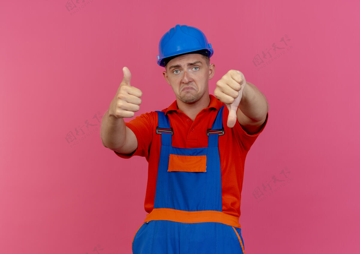 拇指悲伤的年轻男性建筑工人穿着制服和安全帽拇指上下粉红色安全向下头盔