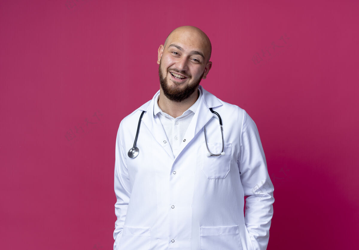 秃头面带微笑的年轻秃头男医生穿着医用长袍和听诊器把手放在口袋里隔离在粉红色的复印空间太空听诊器年轻