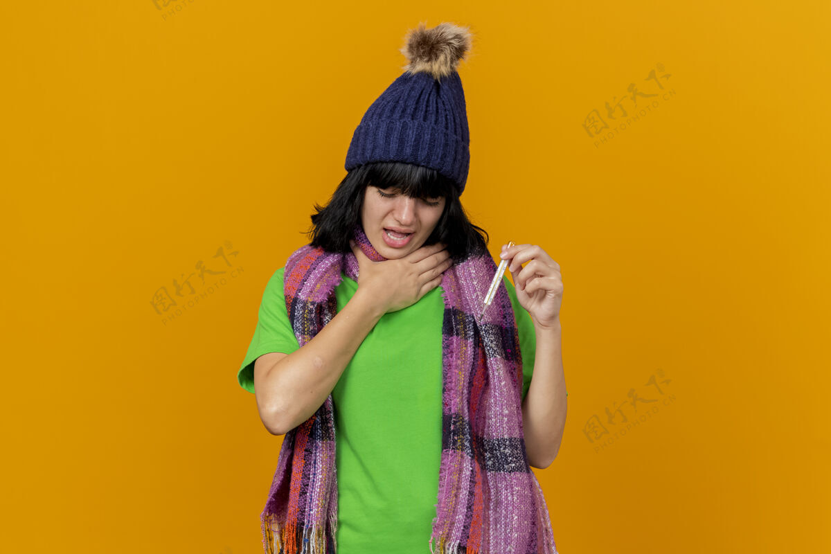 人一个戴着冬日帽子 戴着围巾 手捂着喉咙 闭着眼睛 手拿体温计的年轻生病的女人被隔离在橙色的墙上脸市民疾病