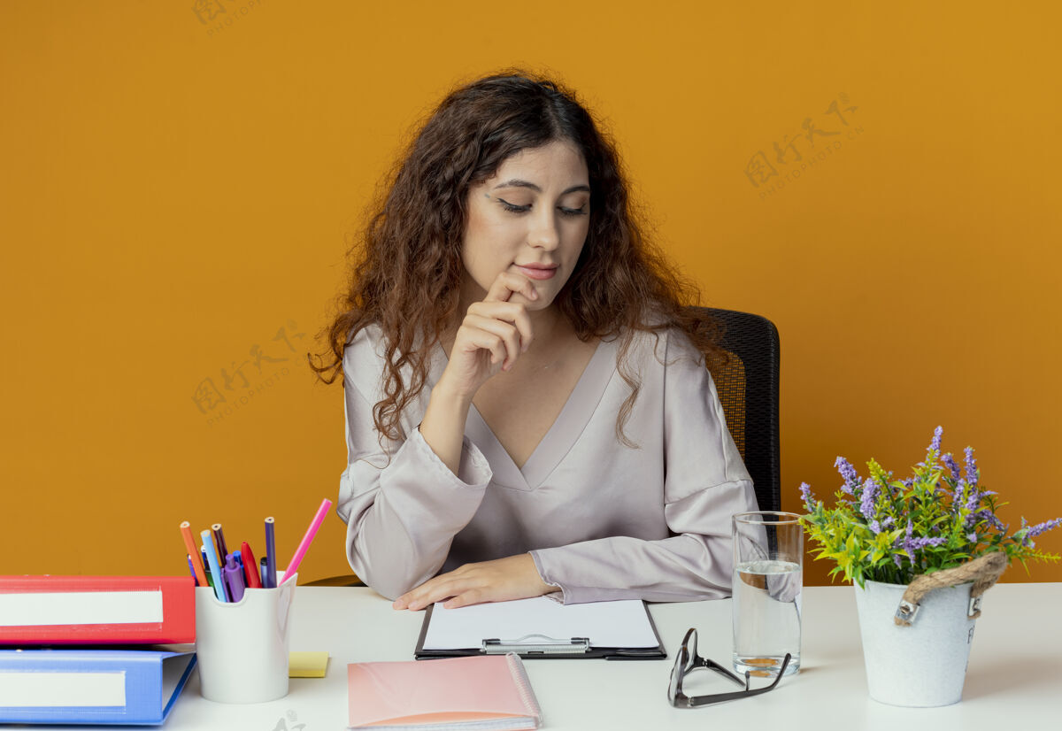墙看着书桌心想：年轻漂亮的女上班族坐在书桌旁 手里拿着办公工具 把手指放在橘黄色的墙上手势年轻漂亮