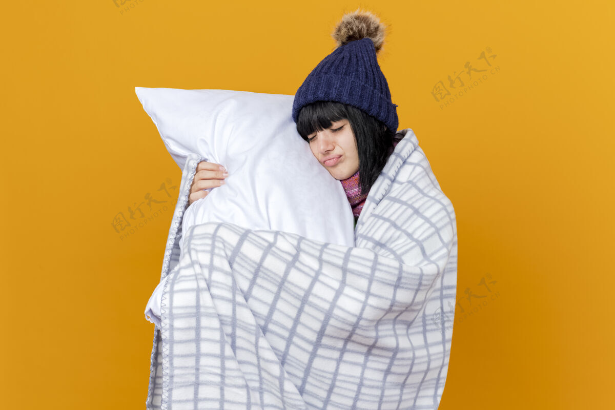 年轻年轻生病的高加索女孩戴着冬天的帽子和围巾 裹着格子布 抱着枕头 用它抚摸着脸 闭着眼睛 在橙色背景上隔离 留着复制空间封闭格子穿