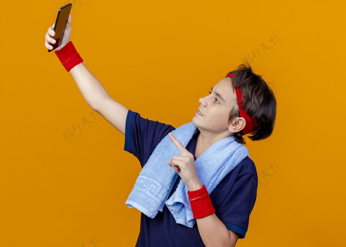 运动年轻英俊的运动男孩戴着头带和护腕 脖子上戴着牙套和毛巾 自拍时指着橘色墙上的手机脸脖子周围