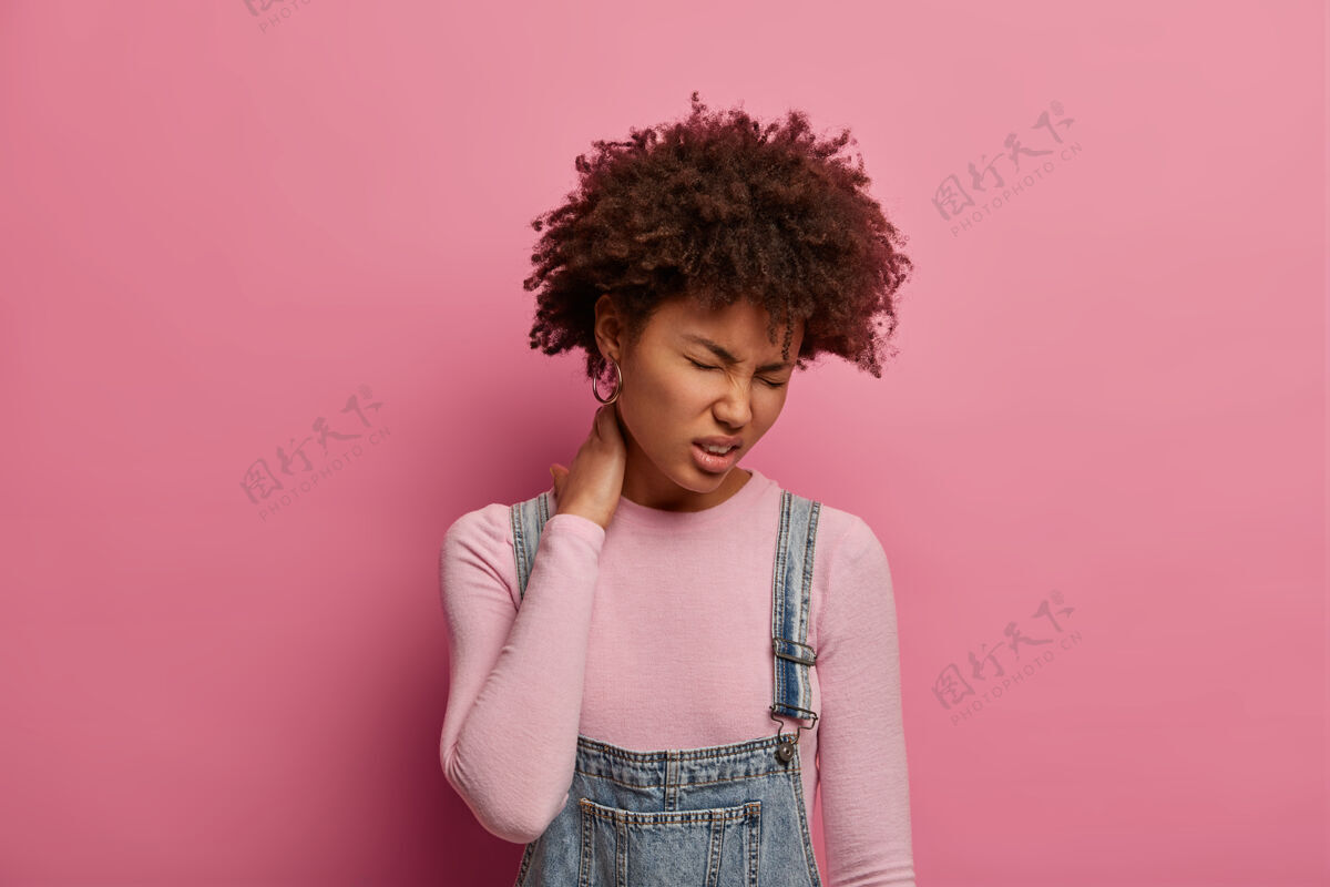 负片不高兴的年轻非洲裔美国妇女感到脊柱不适 触碰颈部和痛苦的皱眉 导致久坐的生活方式 穿着随意 对粉红色粉彩墙姿势 是疲劳颈部感觉受伤