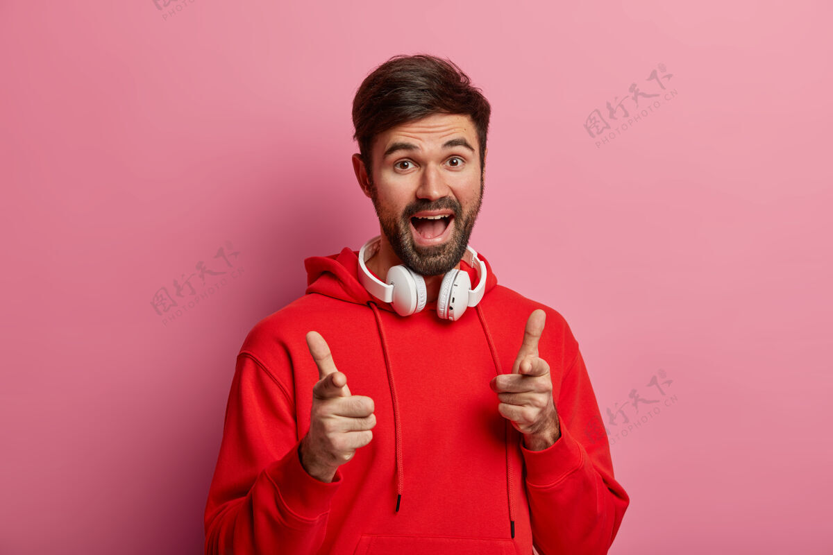 选择积极的留着大胡子的时髦男人直接指着你 做出了很好的选择 说得很好 做出了自己的姿态 戴着红色连帽衫和现代耳机 对着粉色粉彩墙摆姿势请粉彩运动衫