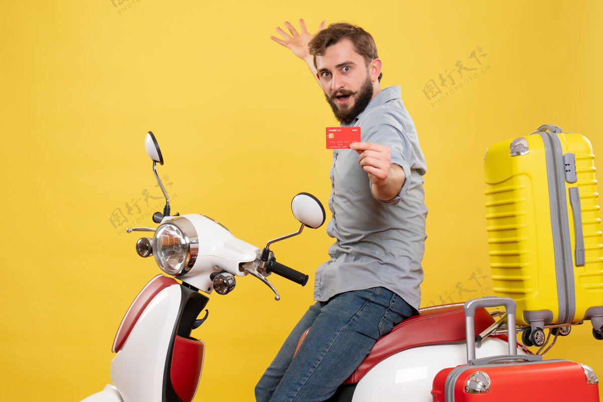 手提箱旅行概念的正面视图 骄傲的雄心勃勃的年轻人坐在摩托车上 手提箱上挂着黄色的银行卡自行车摩托车骄傲