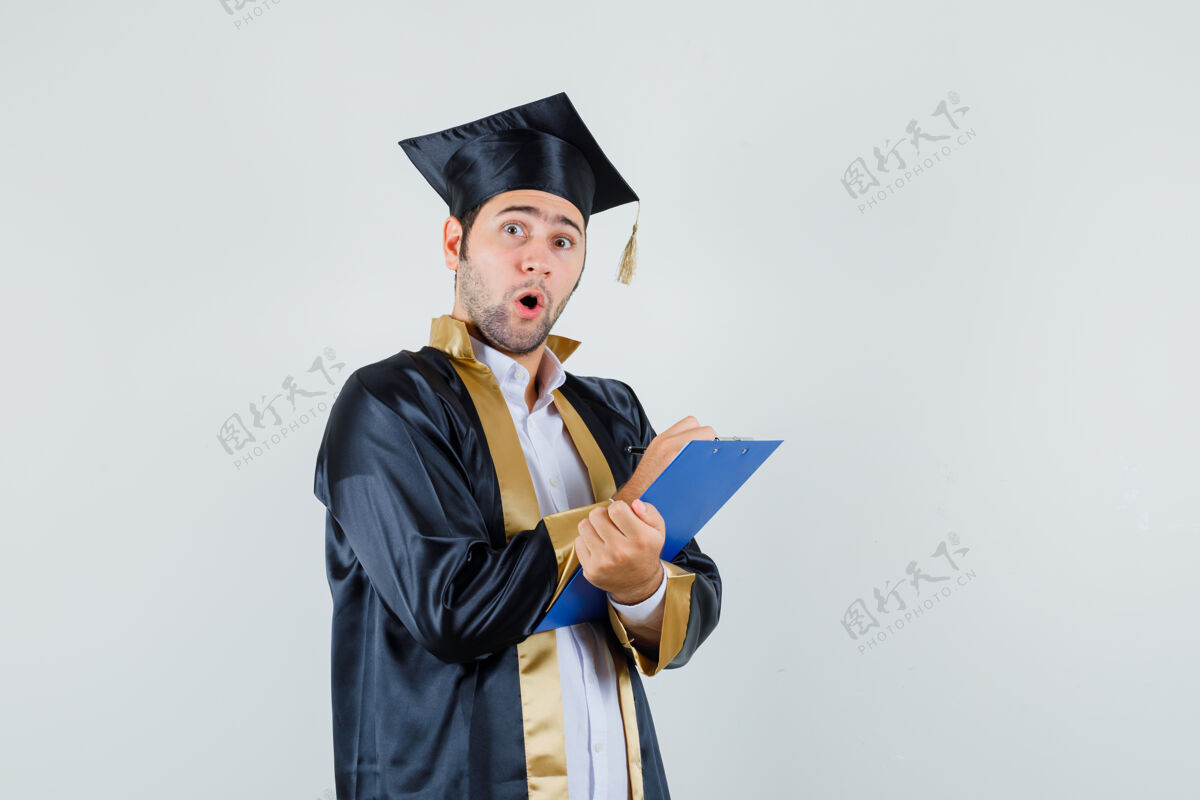 年轻年轻人穿着毕业制服在剪贴板上做笔记 看上去很惊讶前视图英俊成人成功