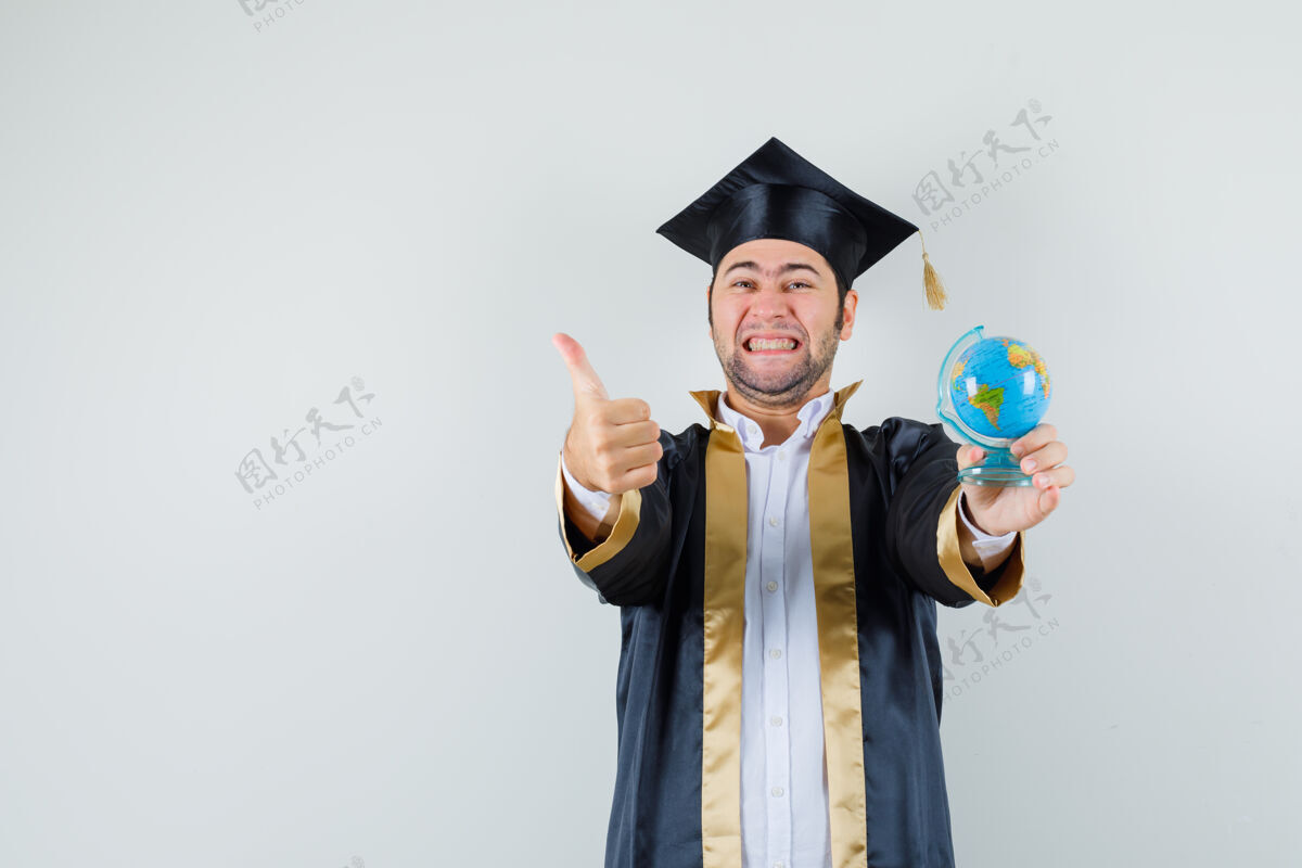 地球仪年轻人拿着学校的地球仪 穿着研究生制服竖起大拇指 看上去很幸运 正前方视图年轻男人肖像