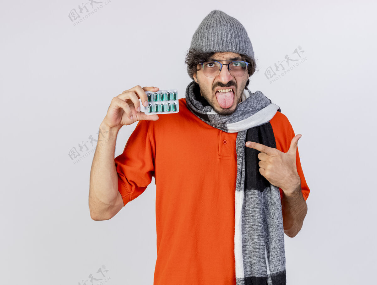 穿着自信的年轻人戴着眼镜 戴着冬天的帽子 戴着围巾 拿着并指着一包医药胶囊 露出舌头 看着隔离在白墙上的前面眼镜衣服年轻人