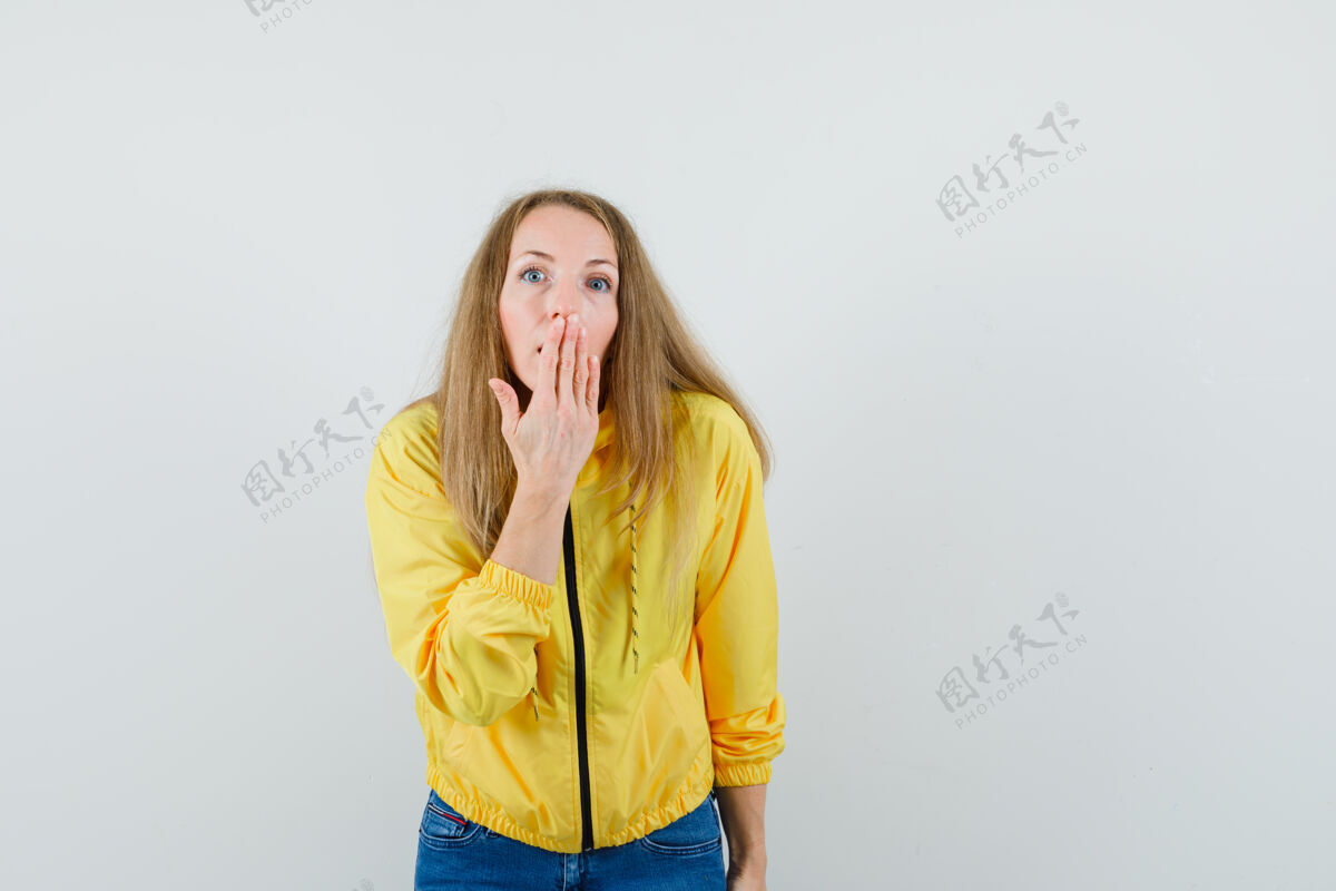 女人身穿黄色棒球服和蓝色牛仔裤的年轻女子用手捂着嘴 看上去很惊讶 正面视图欢呼美女年轻
