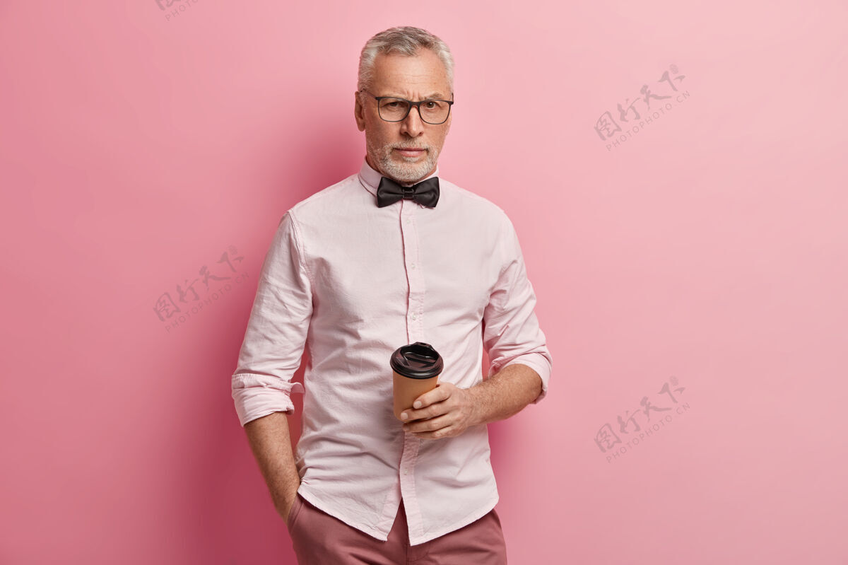 肖像穿粉红色衬衫和黑色领结的老人拿着咖啡杯衬衫人男的