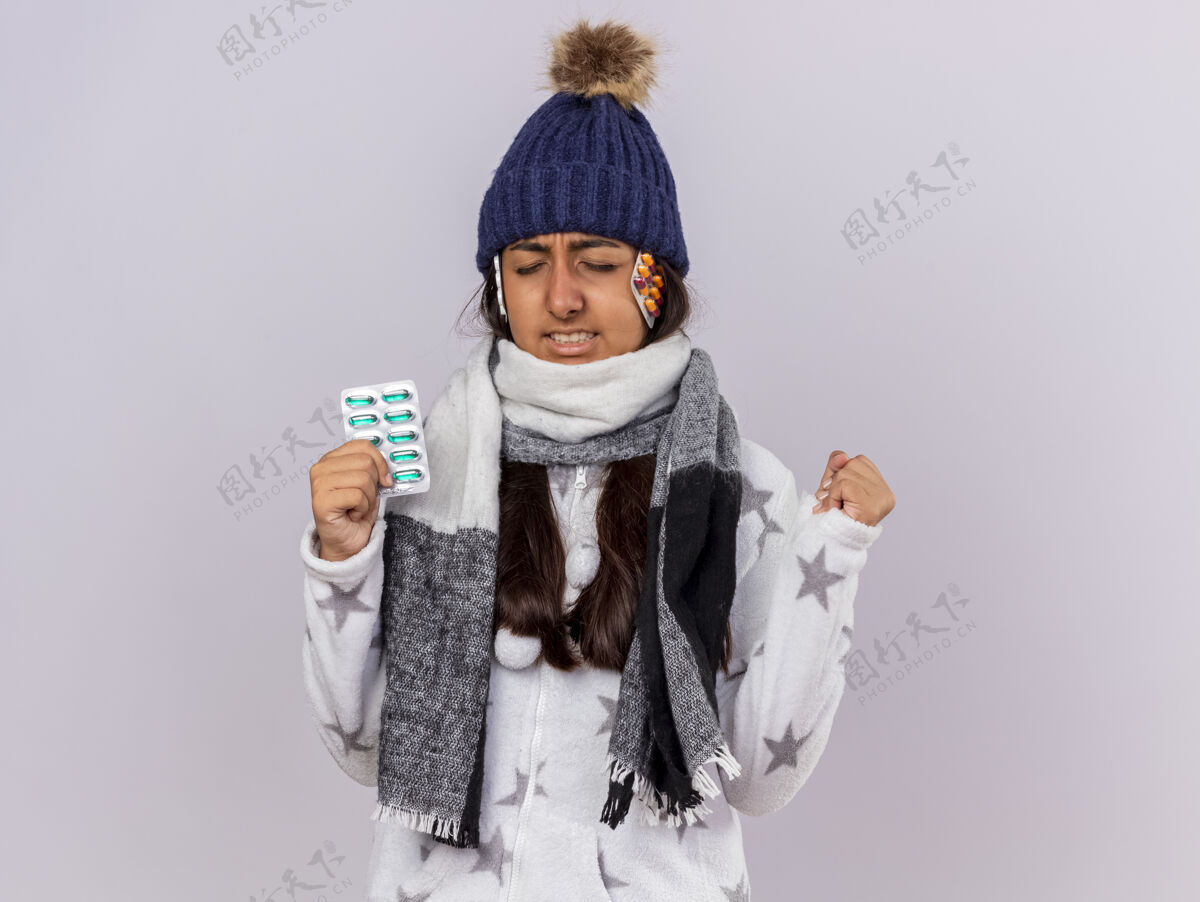 抱着关心的年轻病女孩闭着眼睛戴着冬天的帽子围巾拿着药片把药片放在帽子下面 在白色背景上显示“是”的手势药丸眼睛年轻