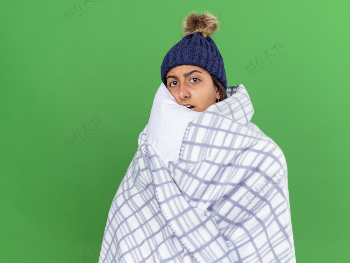 女孩被吓坏了的生病的年轻女孩看着相机 戴着冬天的帽子 围巾裹着格子呢 抱枕孤立在绿色的背景上包装枕头帽子