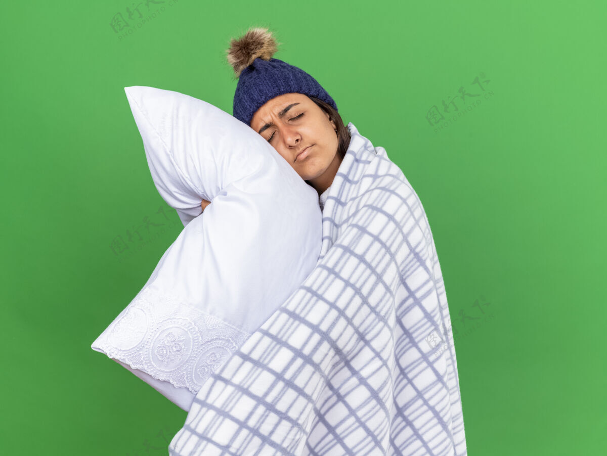 格子布年轻的病女孩闭着眼睛 戴着冬天的帽子 围巾裹在格子布的抱枕里 在绿色的背景下与世隔绝围巾女孩疾病