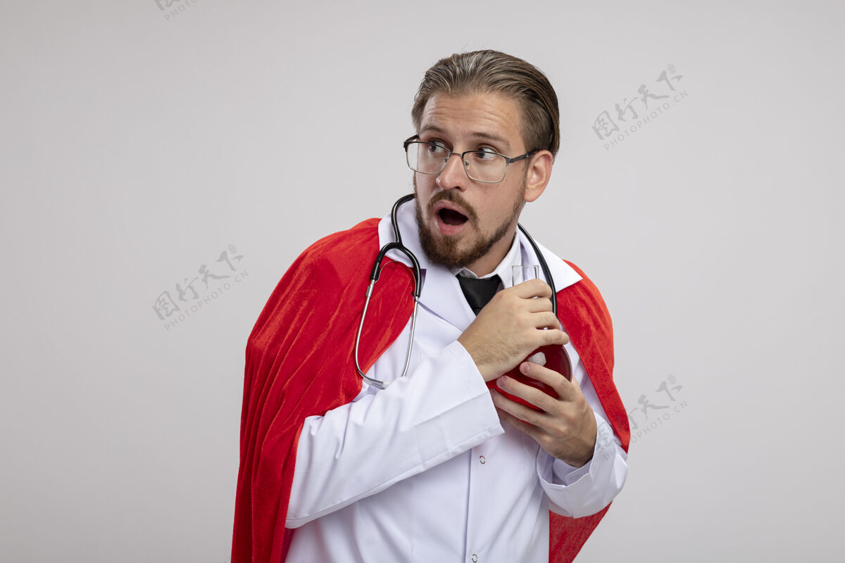 瓶子贪婪的年轻超级英雄 穿着医用长袍 戴着听诊器和眼镜 手里拿着装满红色液体的化学玻璃瓶 隔离在白色背景上年轻液体持有
