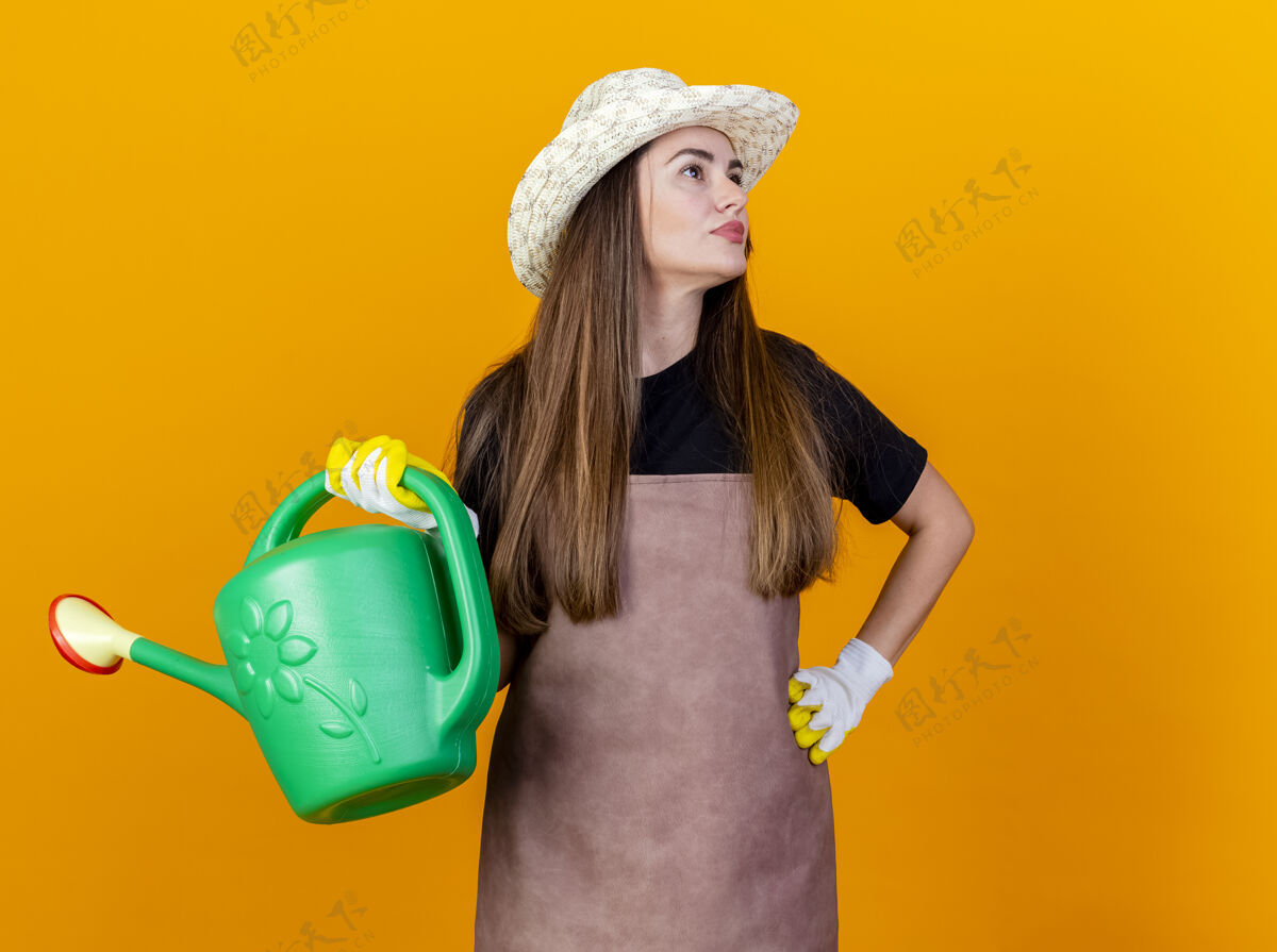 帽子看着身边高兴的漂亮园丁女孩穿着制服 戴着园艺帽 戴着手套 拿着浇水罐 手放在臀部 隔离在橙色的背景上园艺女孩制服