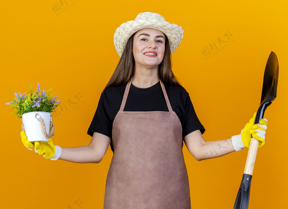 摊开微笑美丽的园丁女孩穿着制服 戴着园艺帽 戴着手套 手里拿着花盆里的花 双手分开放在橙色的背景上黑桃制服手捧