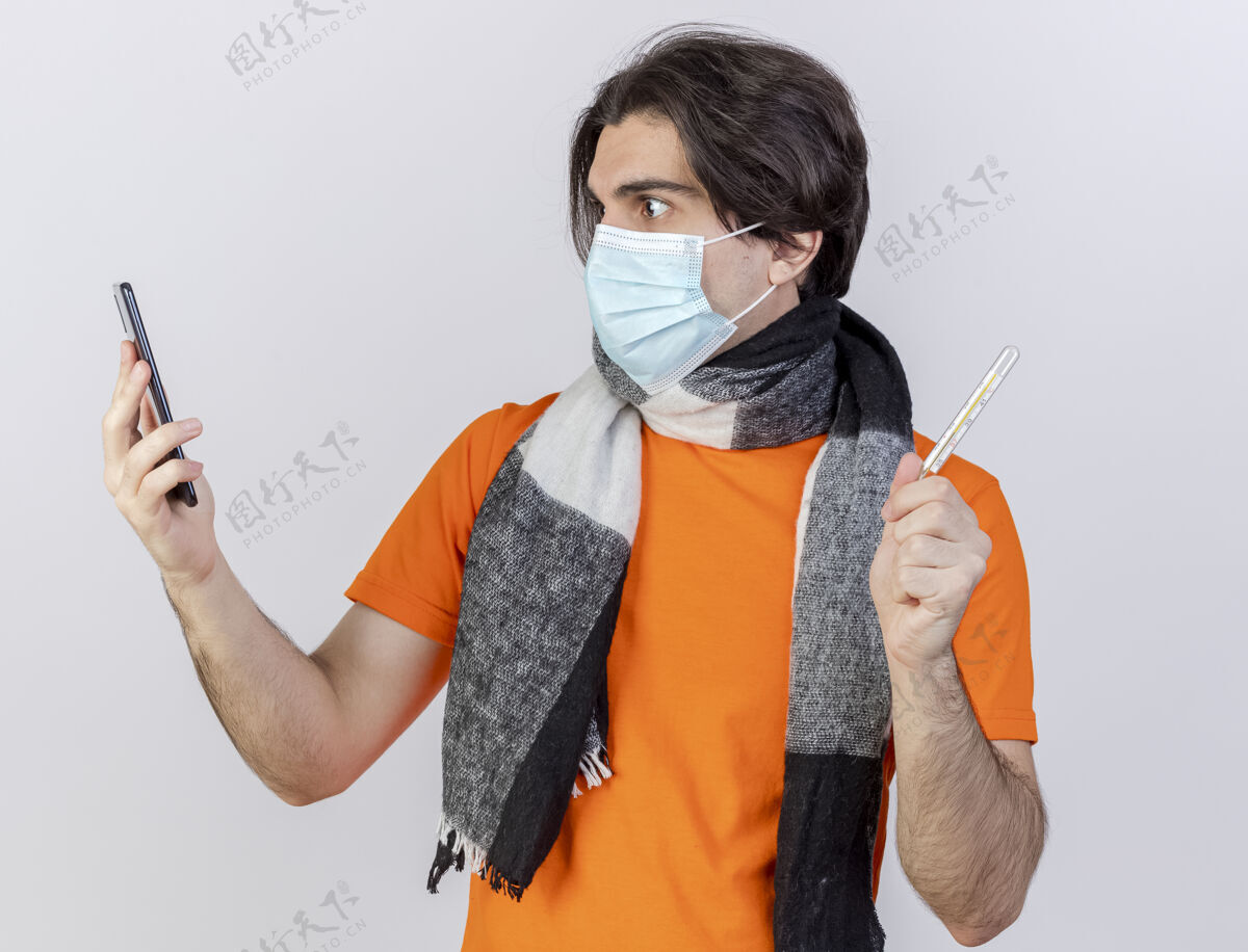 围巾惊讶的年轻病患戴着围巾和医用口罩拿着温度计看着手机在他手上孤立的白色背景医疗疾病面具